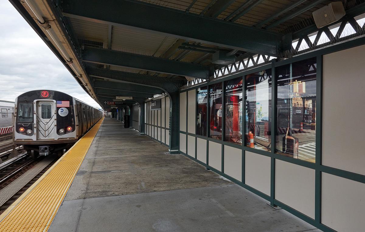 Photograph of laminated glass artwork on the platform walls of the Avenue N station in Brooklyn. The artwork shows photography artwork on glass displaying surface reflections of the street and interiors as well as station reflections, on the right wall. A train pulls into the station on the left.    