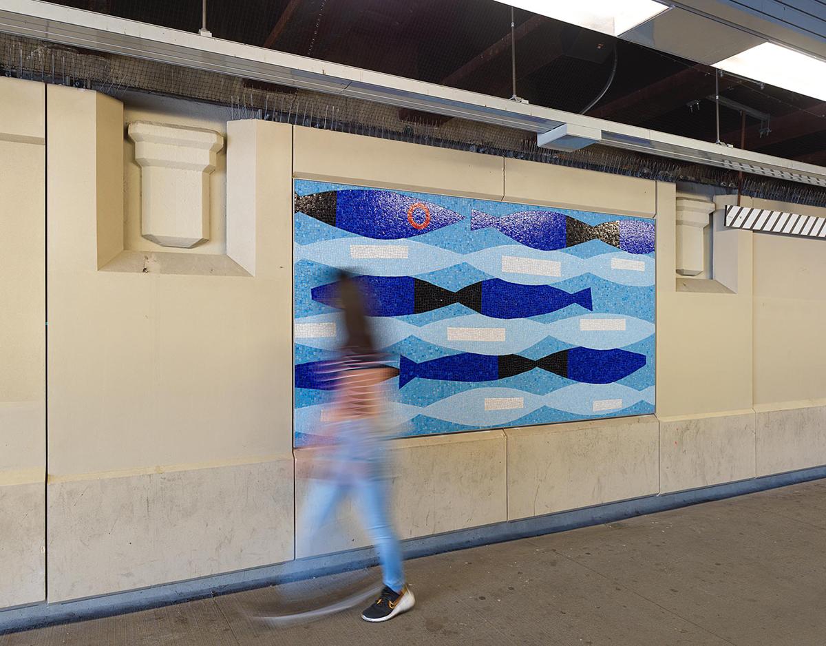 A permanent glass mosaic art installation by artist David Storey on the platform walls of the NYCT 20 Av station shows an abstract composition in shades of blue, reminiscent of a school of fish swimming, with a blurred image of a woman walking in front.