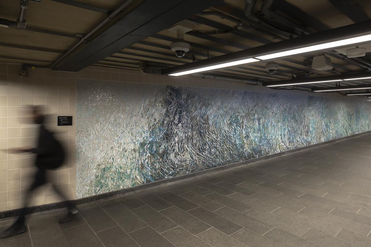 Photograph of mosaic artwork at 34th Street-Penn station corridor left wall. The shimmering glass mosaics in pearls, aquas, greys and gold form white lines that abstractly depict a floor to ceiling ethereal female figure appearing in an atmosphere as a ghostly apparition, with the flowering fabric of her garment stretching the length of the wall. A blurry image of a man walking past the mosaic front is on the left of the photograph and gray floor space is on the right foreground.   