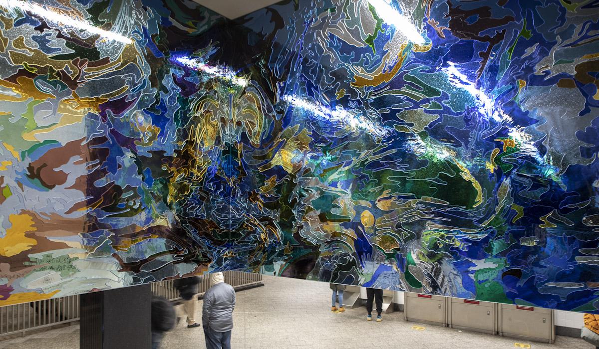 A permanent glass art installation by artist Jim Hodges at NYCT Grand Central Station shows light blue mirrored glass transition to dark blue mirrored glass in camouflage like, organic patterns installed above a stairway with people moving.  