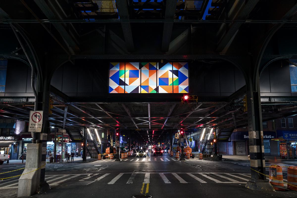Photograph of laminated glass artwork at the 30 Av station in Queens installed along the elevated station’s mezzanine wall, as photographed at night from the street level below. The vibrant color of the large horizontal glass panels of abstract, geometric square patterns in shades of blues and yellows on a white background are illuminated by the surrounding street lights.  