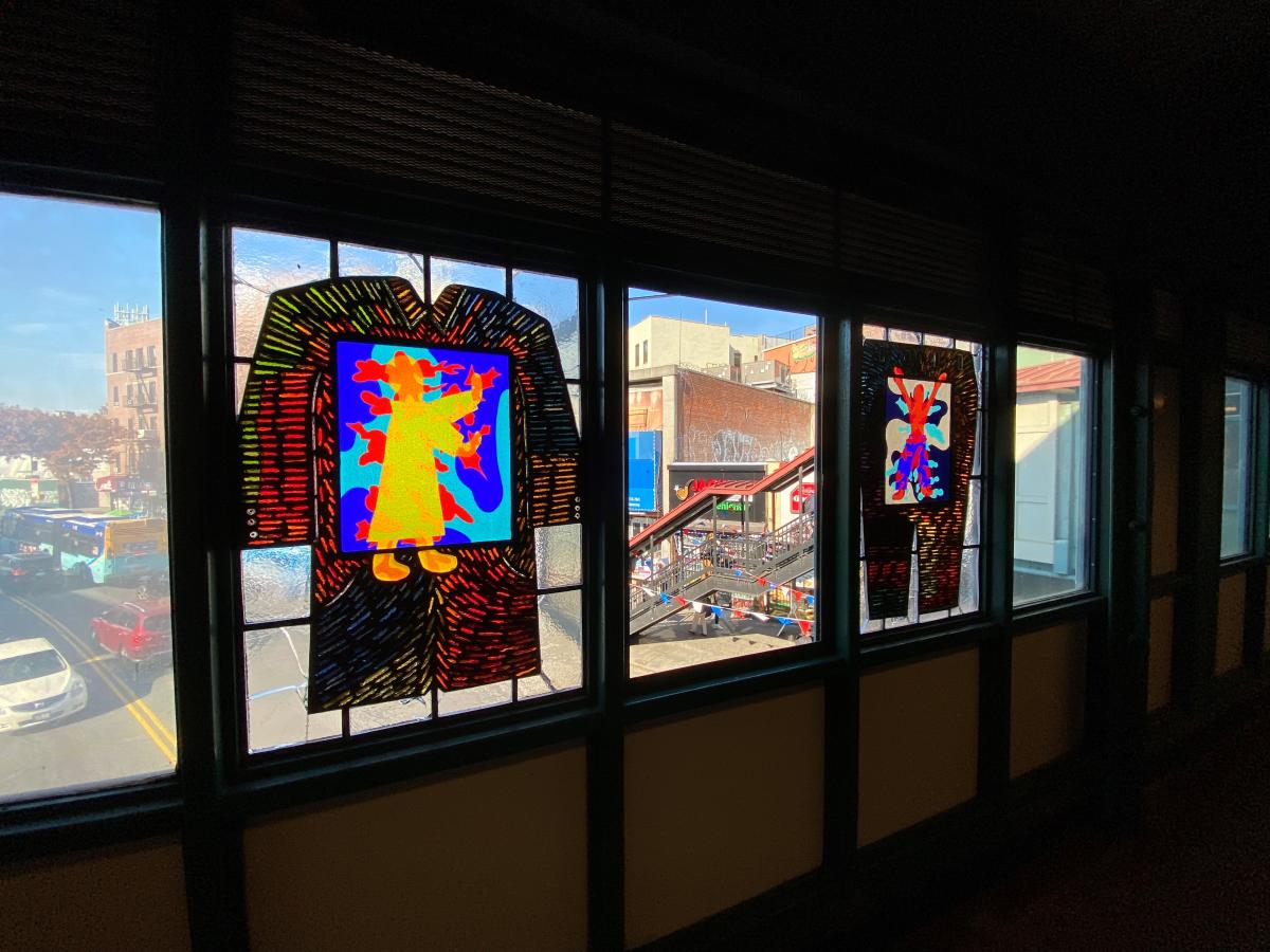 Artwork in faceted/laminated glass by Moses Ros showing colorful figures within backgrounds shaped to represent the items on sale in the surrounding shops. 