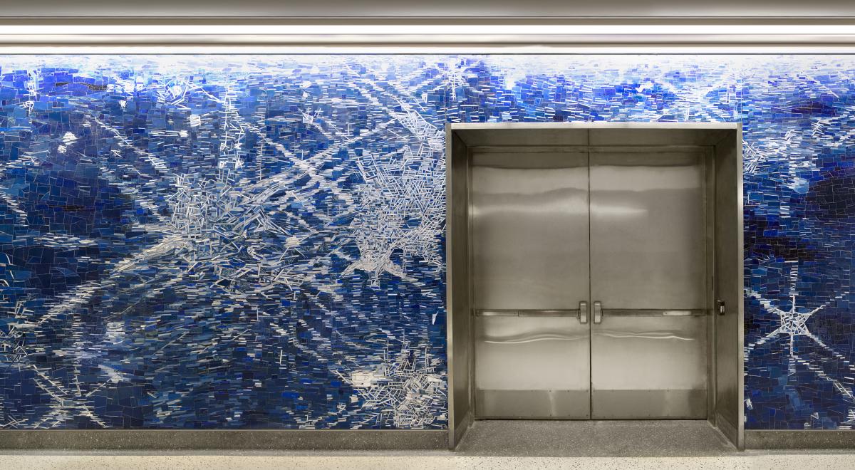 Mosaic artwork along a train station corridor, the mosaic depicts light as it hits a flowing blue river.