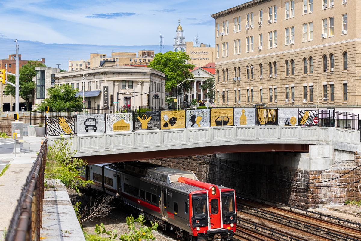 View of a bridge over train tracks. Bridge has yellow, silver, and black metal artwork with icons against patterned backgrounds.