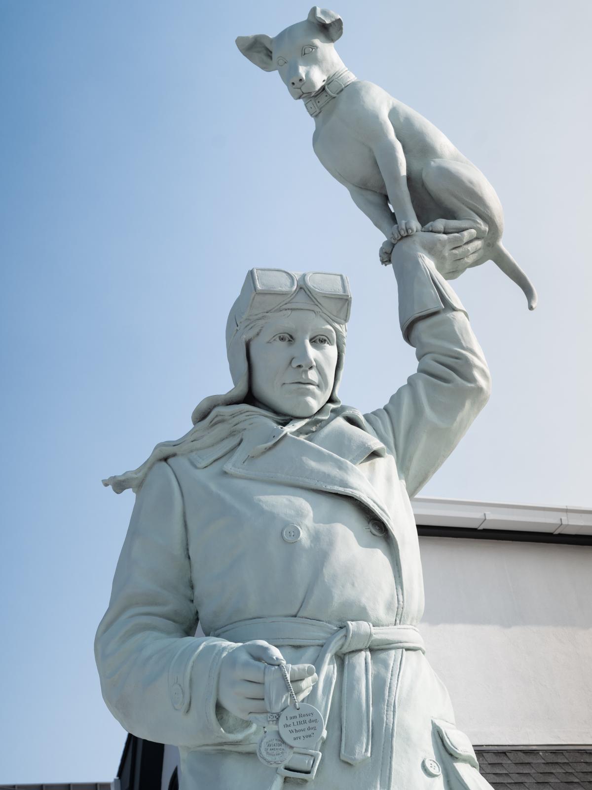 Detail of a tall bronze artwork featuring a woman in an early twentieth century pilot’s uniform holding up a dog with one hand.