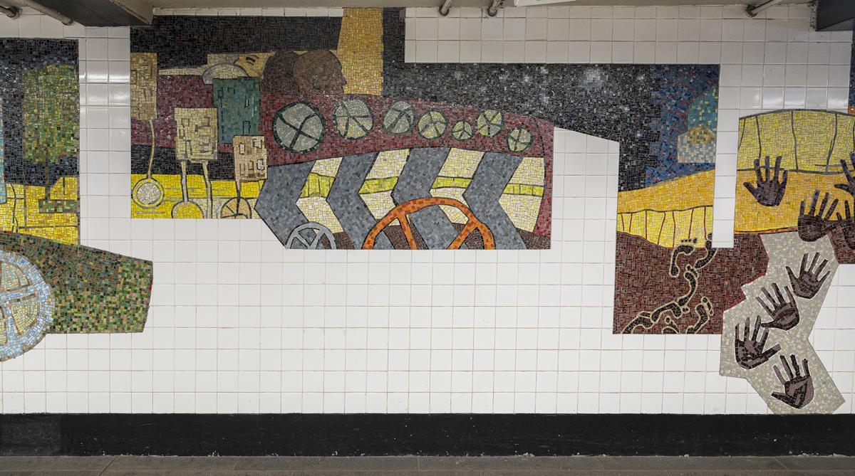 Station hallway with mosaic art on the wall. Mosaic artwork features abstract shapes, handprints, and wheels.