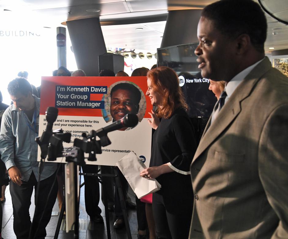 Sarah Meyer, MTA's chief customer officer, smiles near a sign introducing a new group station manager. Microphones and other MTA employees are in the foreground.