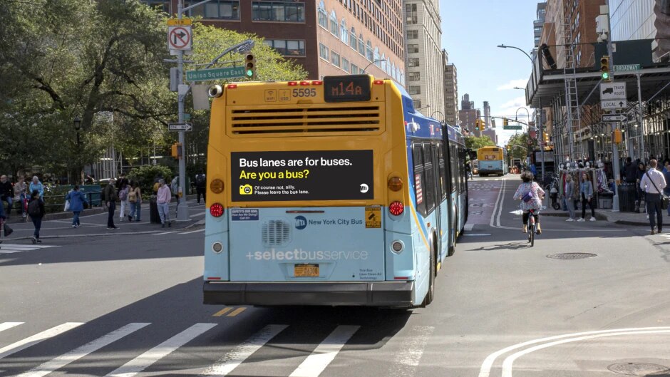 A rendering shows a black sign with white and yellow text on the back of a bus. The sign reads, "Bus lanes are for buses. Are you a bus? Of course not, silly. Please leave the bus lane."