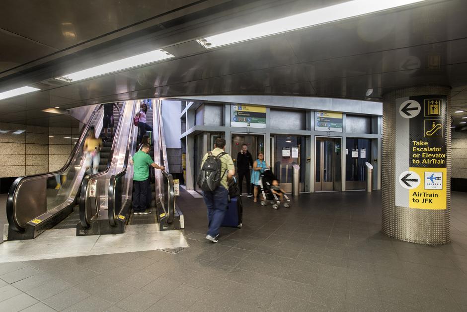 People with luggage take escalators and wait for elevators, with signs on columns nearby pointing to the AirTrain on the upper level. 