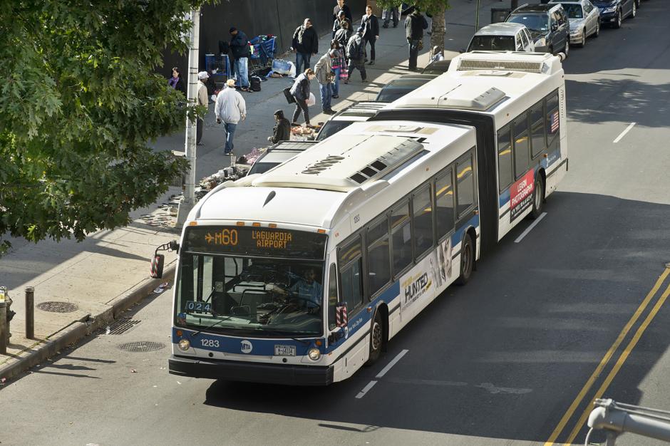 A bus labeled "M60" drives down a city street, with people visible on the sidewalk nearby and sunlight coming from the left side of the frame.