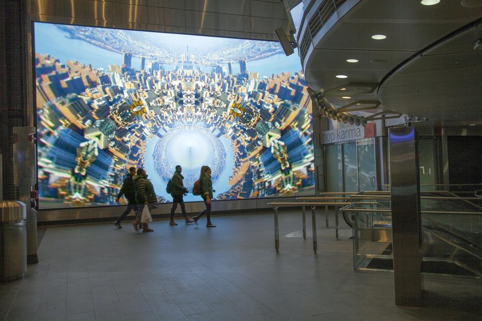 Anne Spalter's digital artwork showing a kaleidoscopic abstraction of the New York City skyline on a large LED screen as four people pass in front of the screen into Fulton Center.