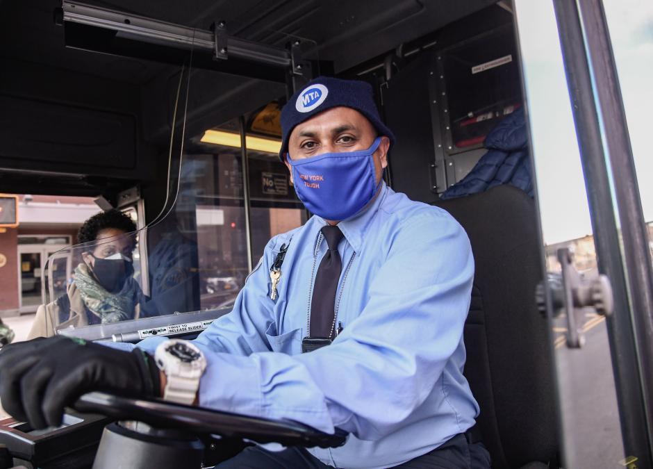 A man wearing a light blue shirt and a dark tie and a face mask behind the wheel of a bus. A woman wearing a coat and a face mask is boarding the bus to his right.
