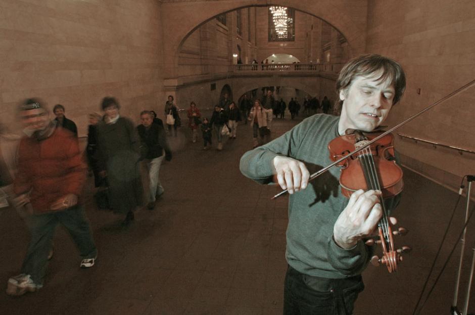 Man playing violin in a corridor with people passing.