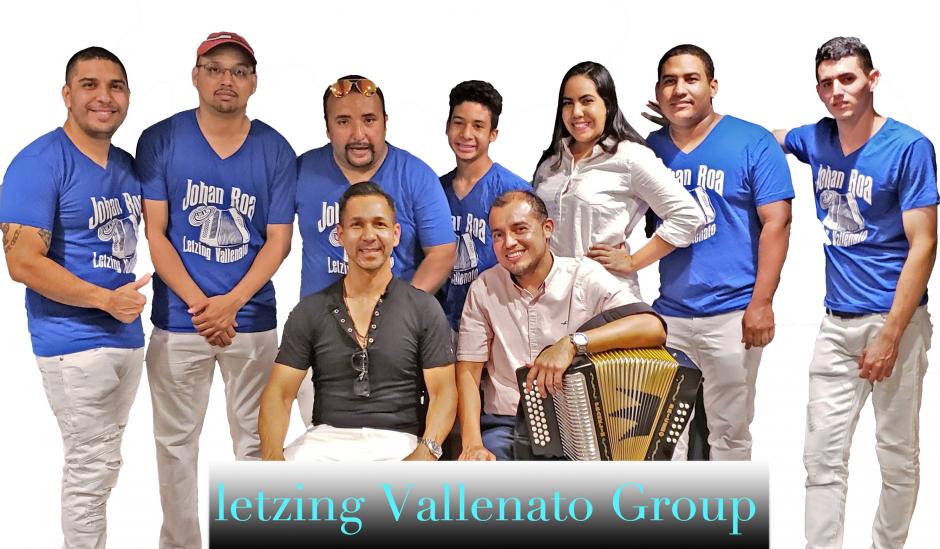 Line of six men posing in matching blue shirts that read Johan Roa and one woman in white blouse with two men sitting in front holding an accordion. A sign at the bottom reads Letzing Vallenato Group.