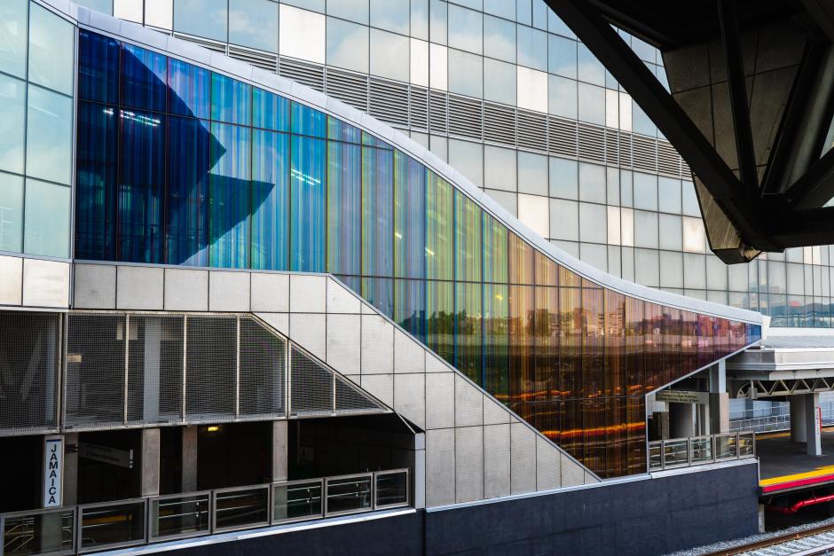 A permanent glass art installation by artist James Little on escalator wall exterior at LIRR Jamaica Station shows colorful intricate lines that together form flat gradated blocks of color in the colors of the rainbow.