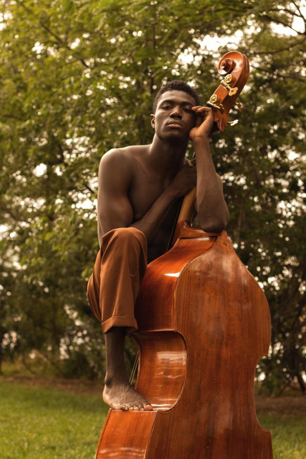 Shirtless man with foot on top of acoustic bass in front of trees.