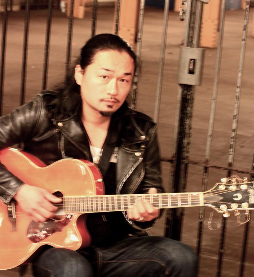 Man in leather jacket playing acoustic guitar.