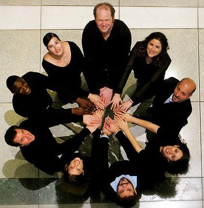 Group of men and women wearing all black standing in circle holding hands in the center looking up at the camera.