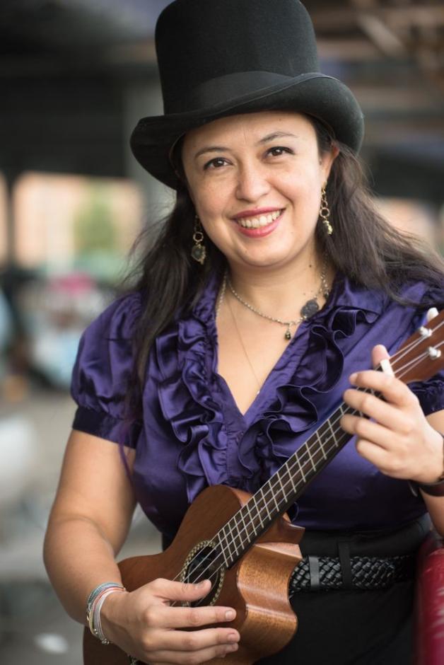 Woman wearing a black top hat and purple blouse holding a ukulele.