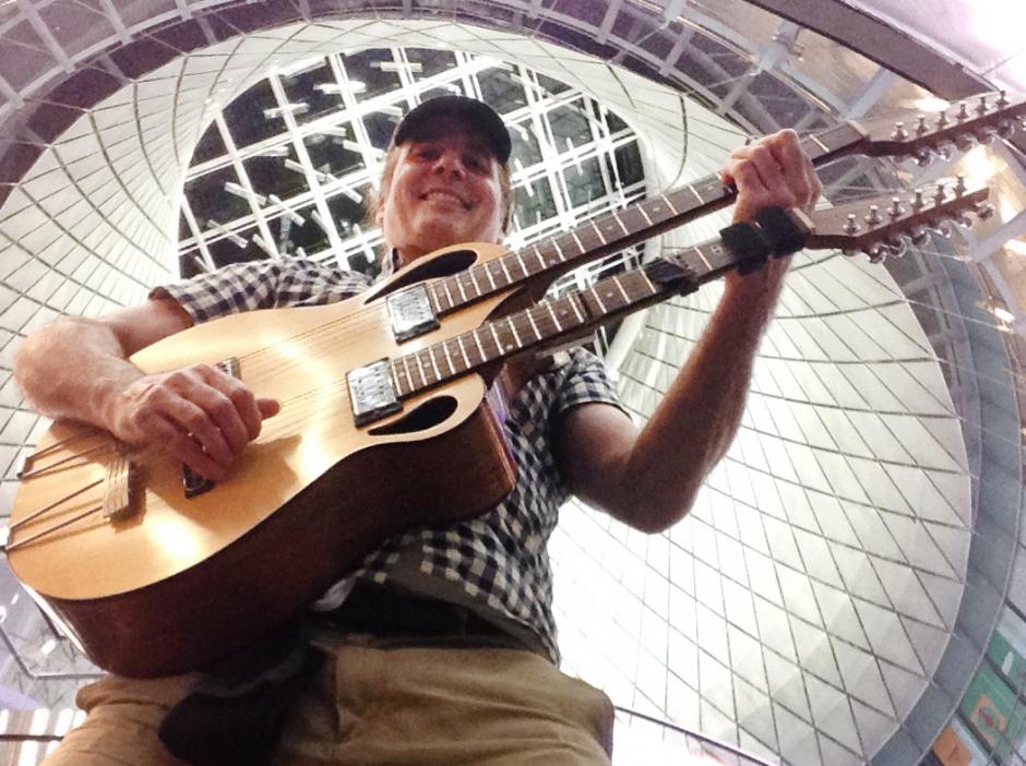 Man playing double neck acoustic guitar In front of architectural patterned background.