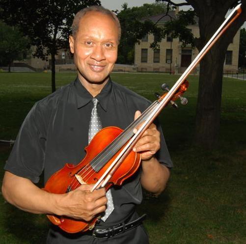 Man in black shirt and tie standing in park holding violin.