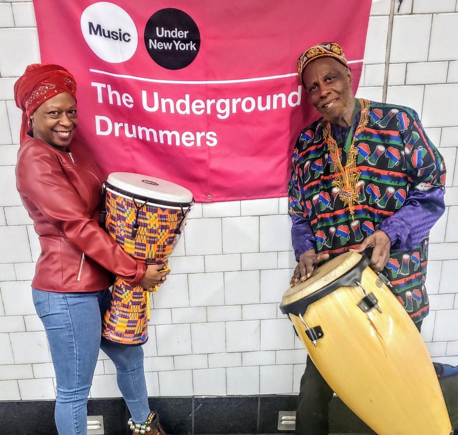 Woman and Man wearing traditional African head pieces holding a djembe and conga drums in front of a sign that reads The Underground Drummers.