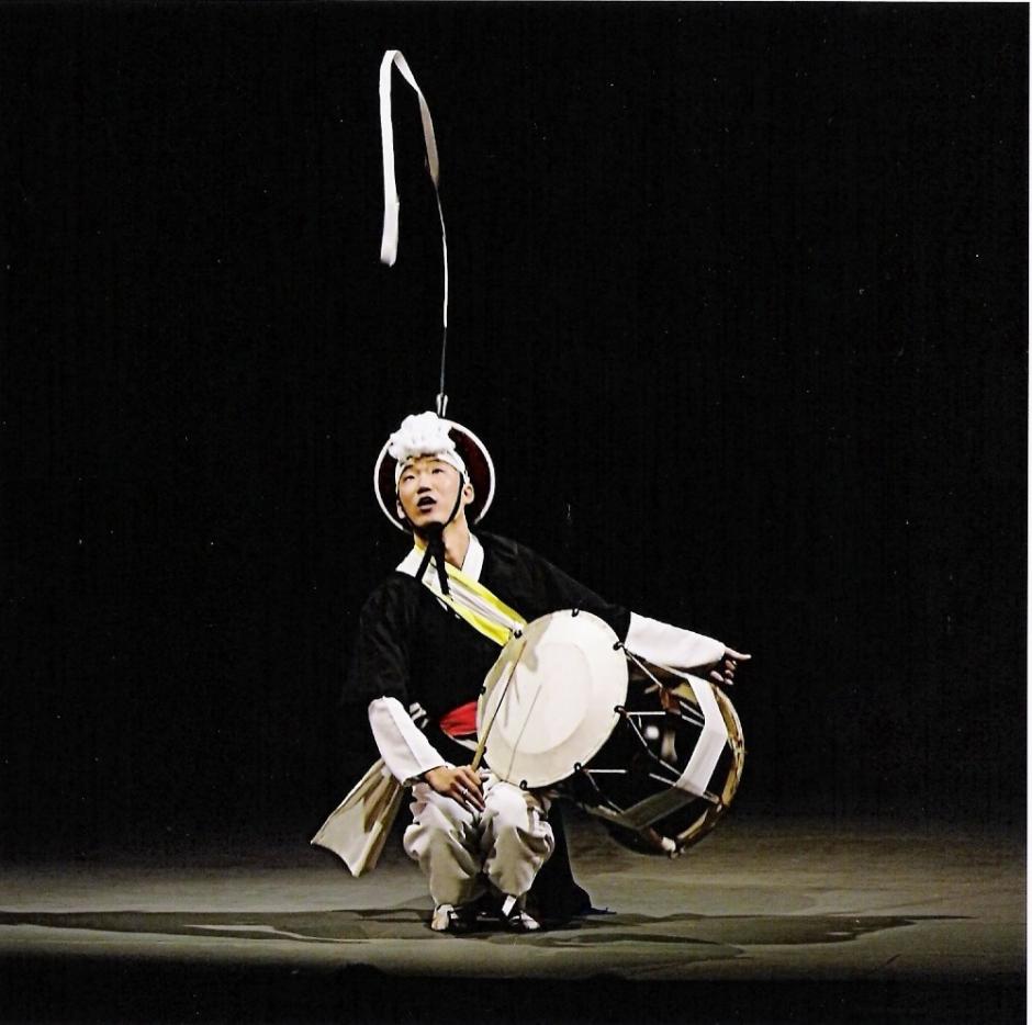 Man in black and white traditional costume playing large Korean drum.