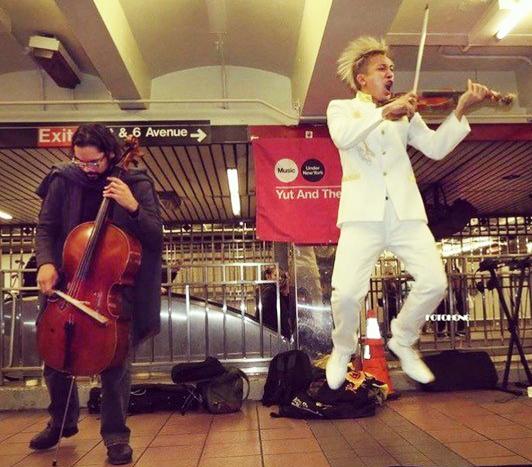 Man dressed in white jumping in the air playing violin and a man dressed in black playing cello.
