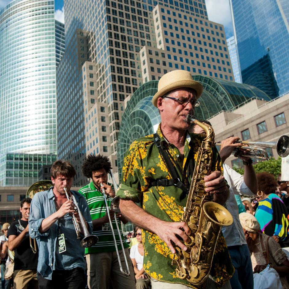 Man in Hawaiian shirt and bowler hat playing alto saxophone with three men playing trombone and trumpet behind. Tall buildings behind them.