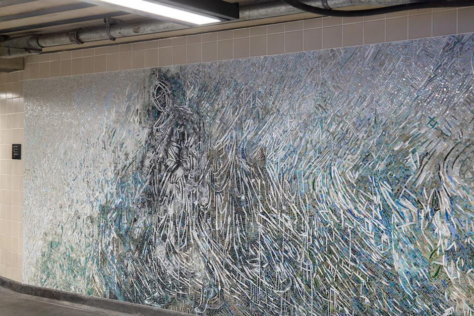 Photograph of a section of the mosaic artwork at 34th Street-Penn station corridor right wall. The shimmering glass mosaics in pearls, aquas, greys and gold form white lines that abstractly depict a floor to ceiling ethereal female figure appearing in an atmosphere as a ghostly apparition, with the flowering fabric of her garment stretching the length of the wall.
