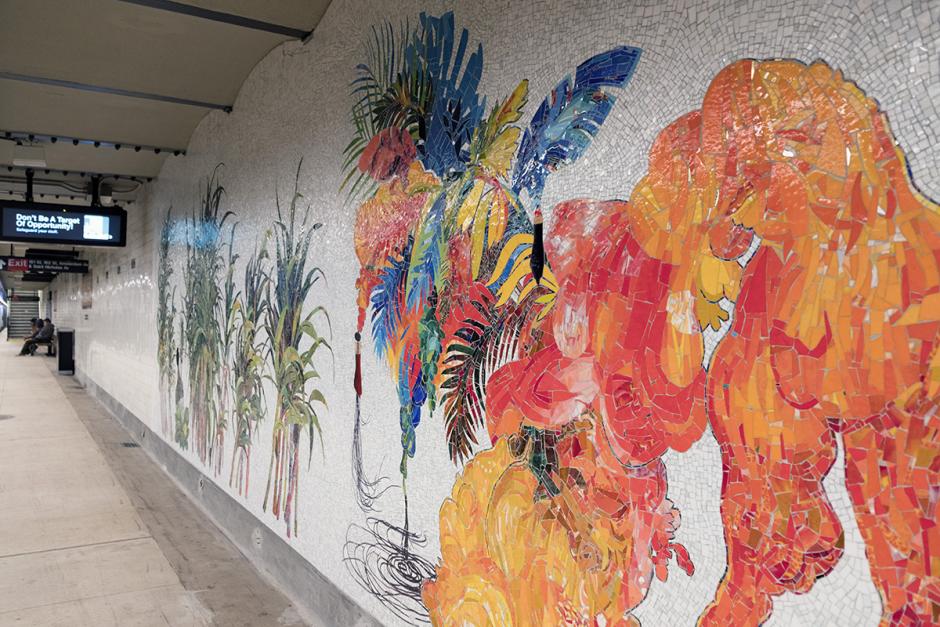 Angled photograph of Firelei Báez’s mosaic on station’s platform wall. The photo shows a ciguapa with orange mosaic tiles next to blue, orange and red leaves and feathers.
