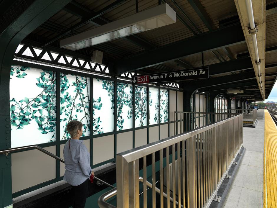 Photograph of the Bay Parkway station platform, with an angled view of laminated glass artwork panels installed on the station platform. Panels feature branches with green leaves on a white background.