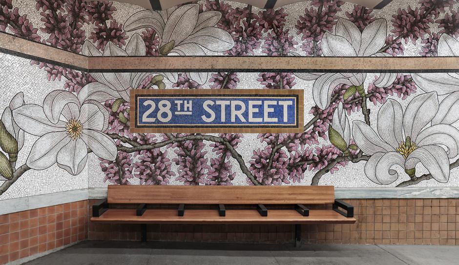 A permanent glass mosaic art installation by artist Nancy Blum on the platform walls of the NYCT 28 St station shows pink and white flowers covering the station walls and wrapping around the mosaic station signage, with a wooden bench in the foreground.
