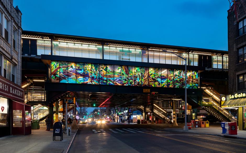 Photo of station exterior at night with Diane Carr laminated glass artwork on mezzanine level in center of image. The artwork is of a forest scene with blues, purples, browns and greens.
