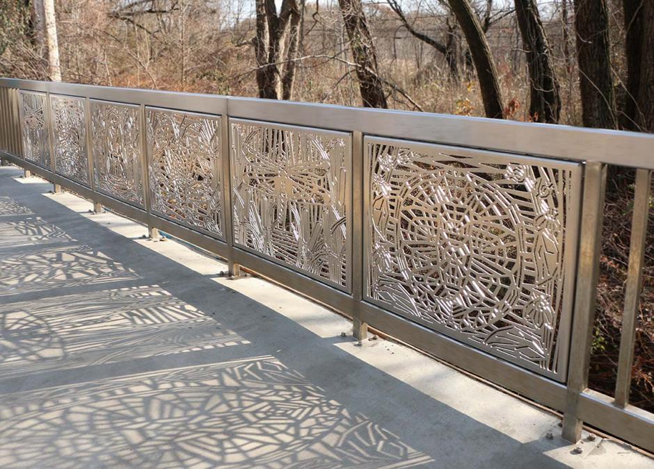 Photograph of metal artwork at SIR Richmond Valley station. The series of stainless steel artwork panels along the station railing depict a spiderweb and dragonfly in the foreground, and the sunlit fence casts a shadow of lines across the station platform. 