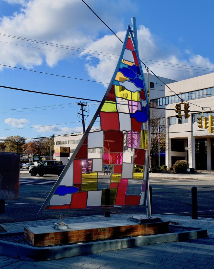A permanent stainless steel and plexiglass sculpture by artist Tom Fruin at LIRR Syosset station shows a tall sail on a concrete base with blue sky and a traffic light visible through a patchwork of plexiglass colors.  