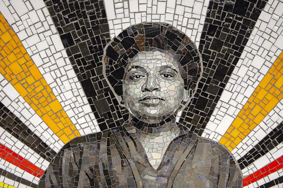 A close-up photograph of a mosaic featuring Audre Lorde with black, red, and orange stripes shining out behind the figure. 