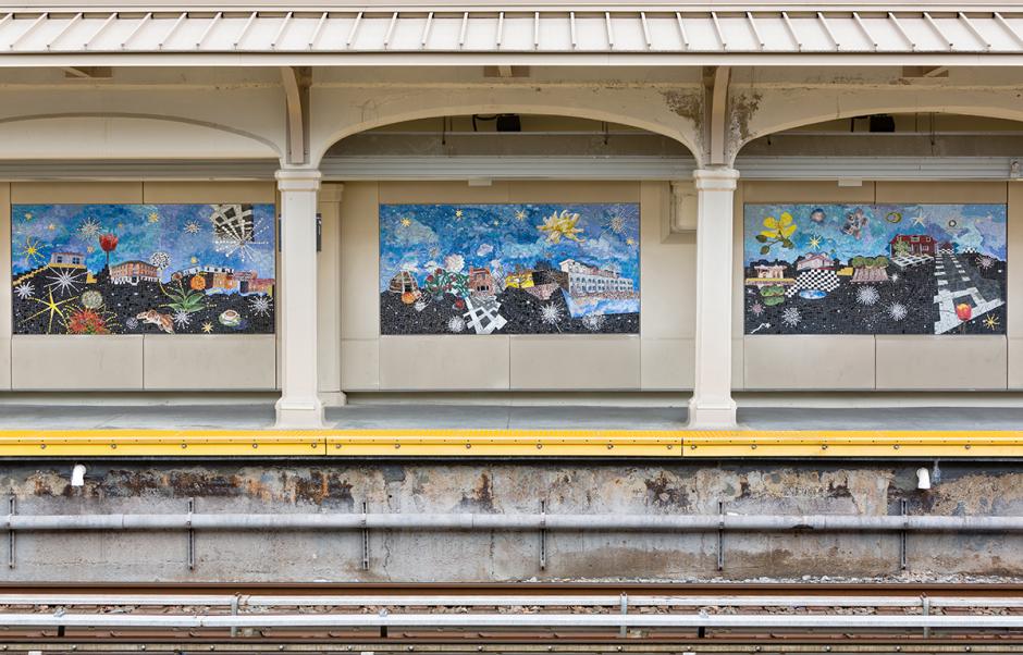 Photograph of mosaic artworks installed on the platform wall of the Avenue U station. The mosaic panels with blue skies and black grounds with objects floating around the horizon line are visible on the platform walls as seen from across the platform, with the yellow platform edge visible below.
