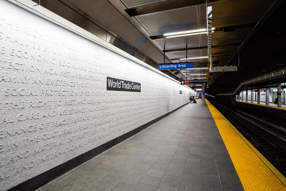 Photograph of marble mosaic artwork on the platform walls of the WTC Cortlandt station.  The white platform walls are lined with words taken from the U.S. Declaration of Independence and the 1948 United Nations Universal Declaration of Human Rights forming a monochromatic tactile surface on the left wall, with a large black and white station name sign sitting on the wall surface, and the yellow platform edge stripe runs down the right side. 