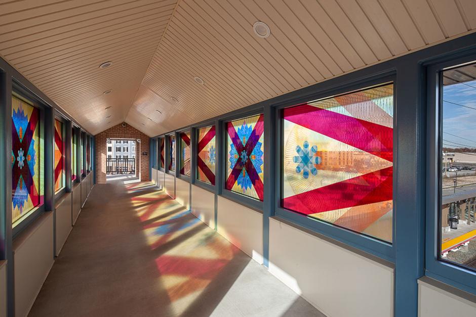 Photo of interior passageway with colorful glass artwork by Mary Judge in windows on both sides. The colorful glass is reflecting color on the ground.