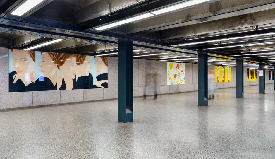 Photograph of laminated glass artworks on wall of 57th Street station. Five hand painted colorful glass artworks, portraits and flowers, line the stone walls of the station, with columns running down the middle of the mezzanine floor and blurred people walk past the artworks. 