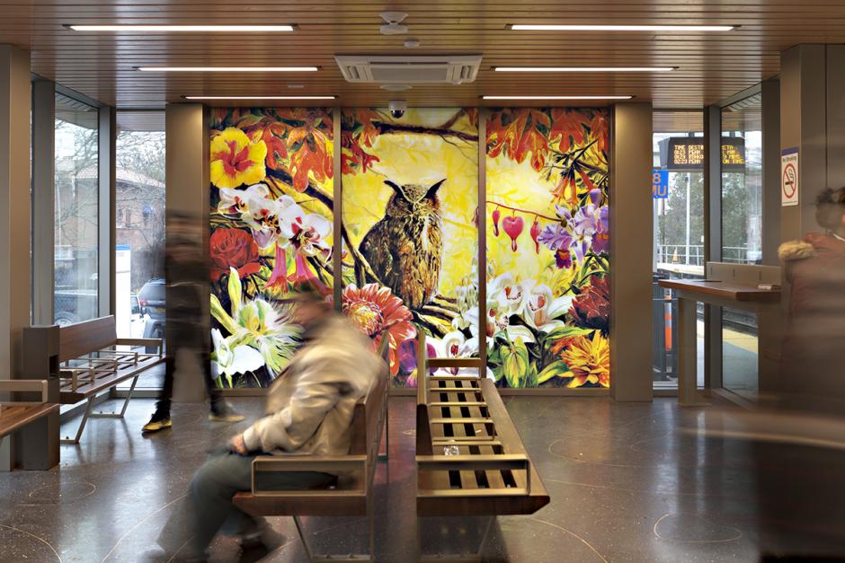 The photo features a station interior view with people seated in a seating area and glass artwork. The glass artwork shows a large owl sitting amongst flowers and branches on top of a yellow background. 
