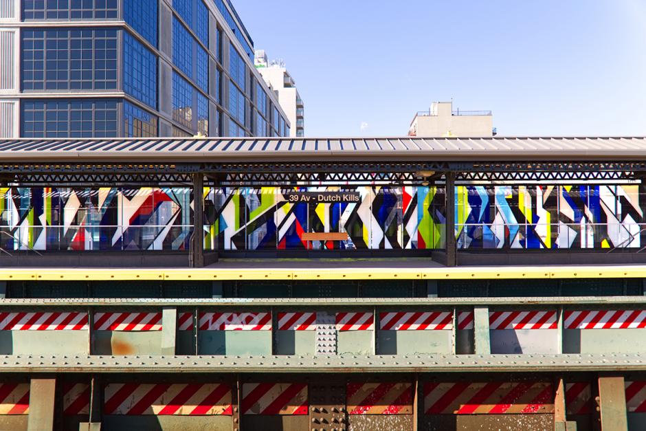 A photograph featuring an across the platform view of glass artwork wall of 39 Av – Dutch Kills Station. The glass artwork is comprised of geometric forms in the colors of red, white, blue, yellow and black. 