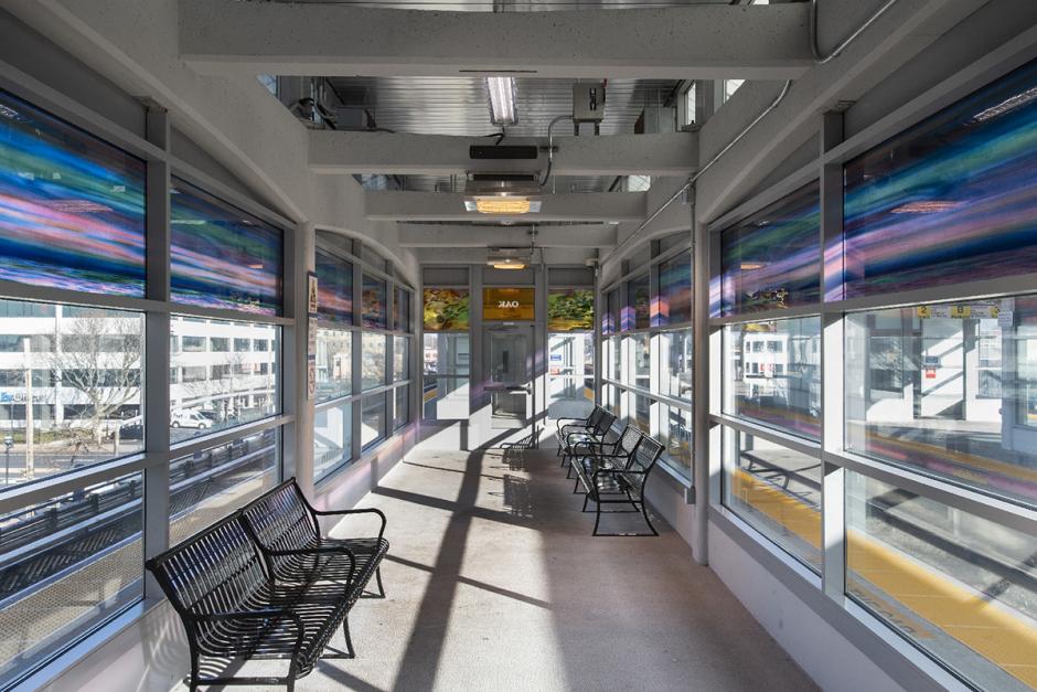 Photograph of laminated glass artwork in the LIRR Hicksville station waiting room. The artwork’s bold colors of purples and pinks line the upper part of the waiting room walls, and a sign above the door read Oak from the reverse. The benches in the waiting area are empty.