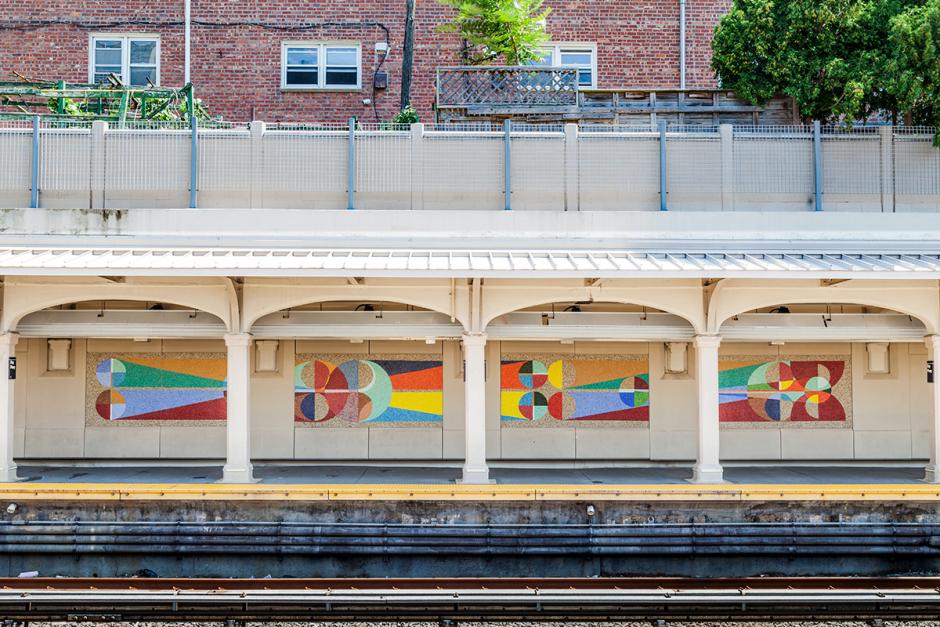 A photograph of glass mosaic artwork on the platform walls of the Bay Parkway station in Brooklyn. Four panels of the colorful geometric abstract artwork are seen from across the platform with tracks in the foreground and brick buildings outside of the station in the background. 