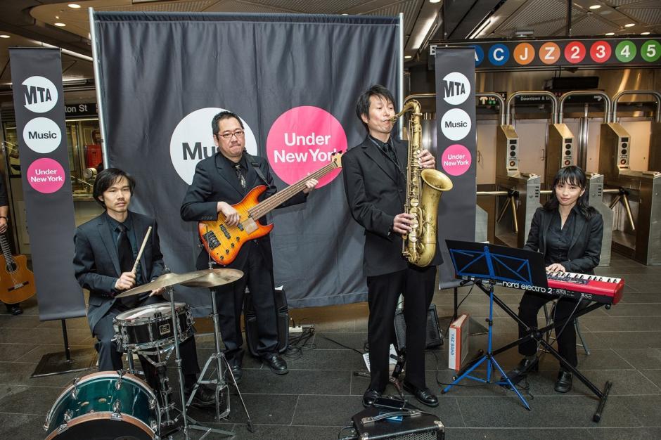 Three men playing Bass, Drums, Saxophone and one woman playing keyboards in front of sing reading “Music Under New York.”