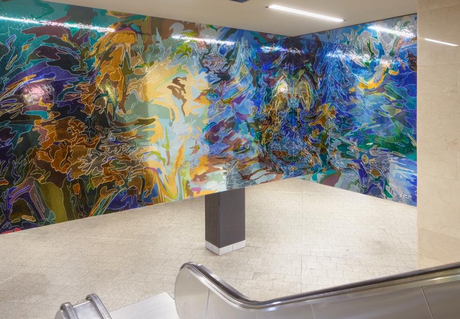 A permanent glass art installation by artist Jim Hodges at NYCT Grand Central Station shows light blue mirrored glass transition to dark blue mirrored glass in camouflage like, organic patterns installed above a stairway.  