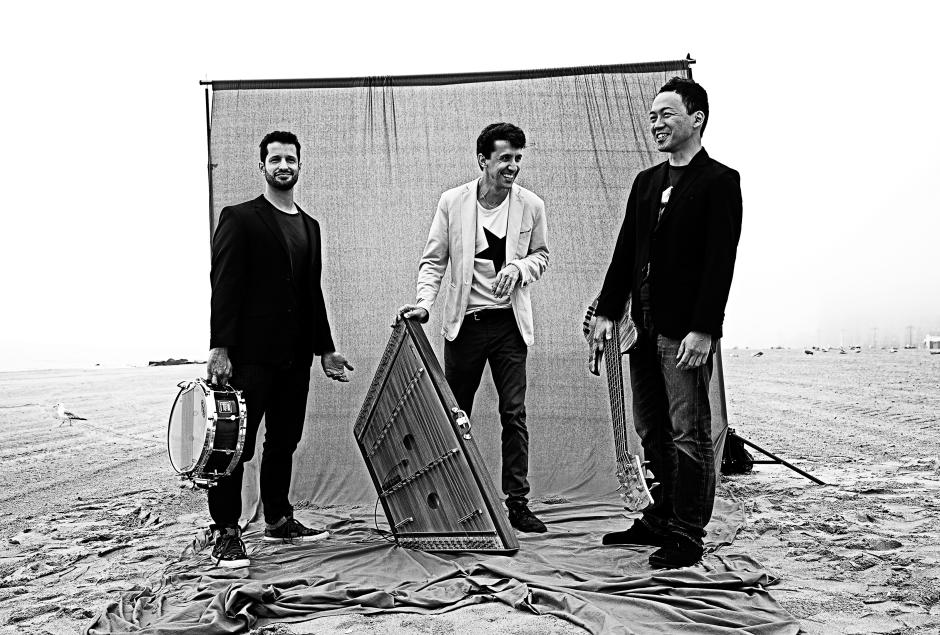 Three men in front of screen on the beach holding a snare drum, hammered dulcimer, and electric bass.