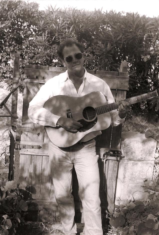 Black and white photo of man playing acoustic guitar in front of a gate.