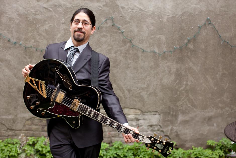 Man in a grey suit posing with an electric/acoustic guitar in front of a wall.